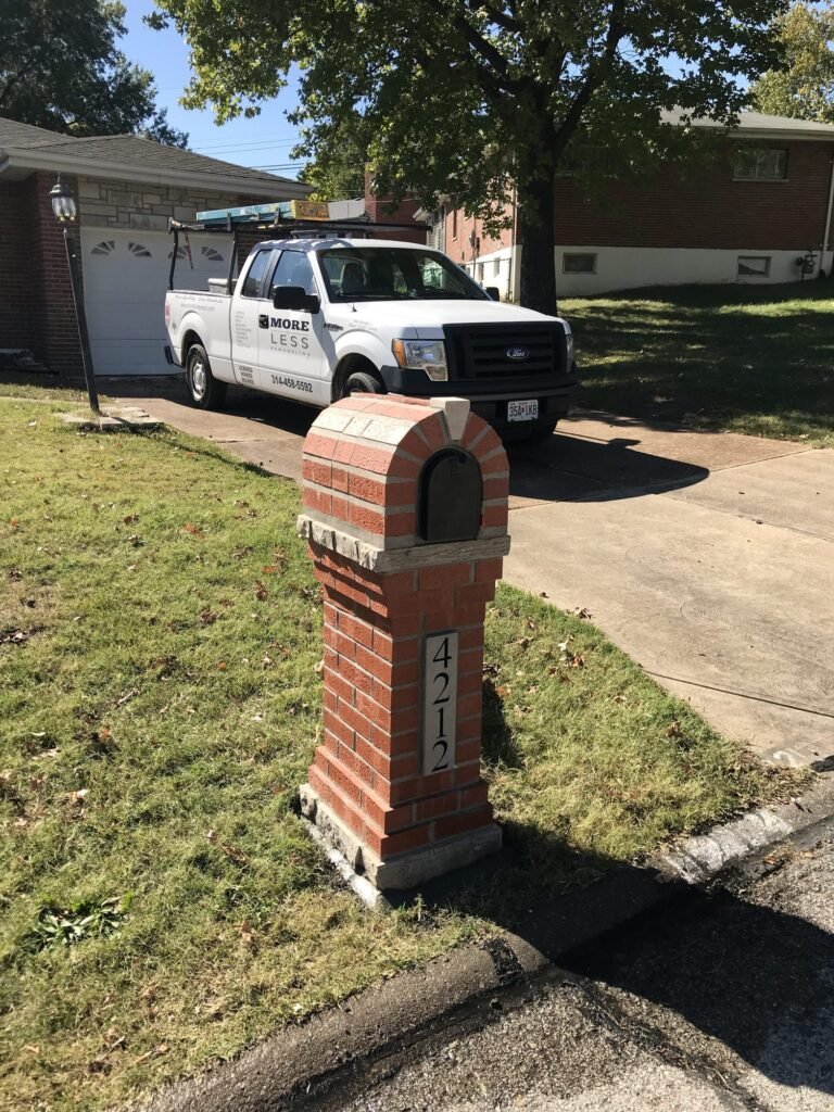 Arched red brick mailbox on a stone cap beside a driveway with a parked work truck