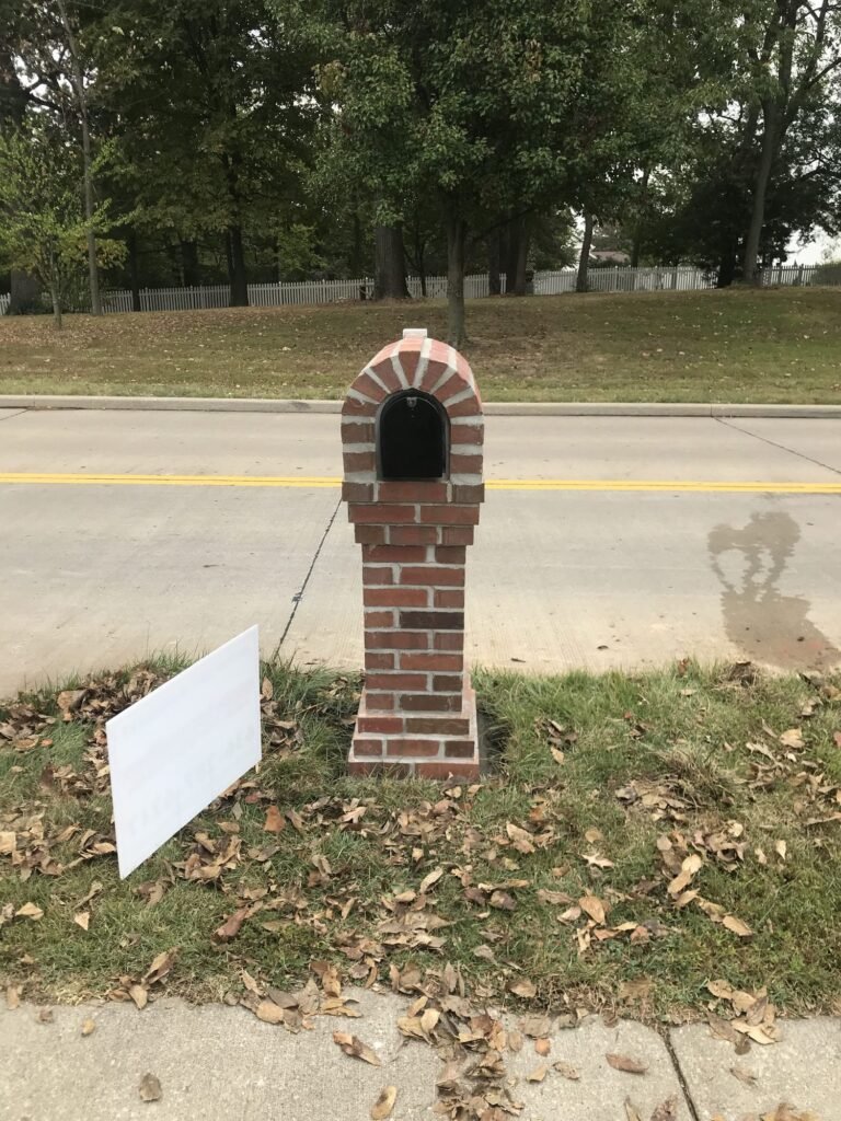 Arched brick mailbox on a grassy median across a two-lane street, fallen leaves around the base