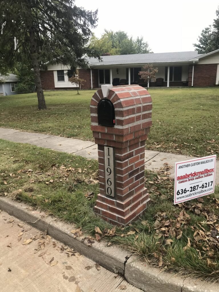 Arched brick mailbox with a builder's yard sign nearby, sidewalk and ranch house in the background