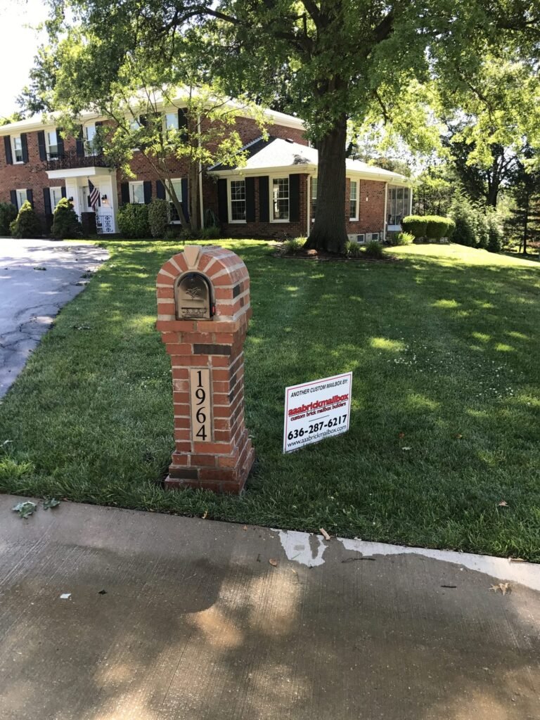 Arched brick mailbox on a tapered stone base, shaded by a mature oak, two-story brick house behind