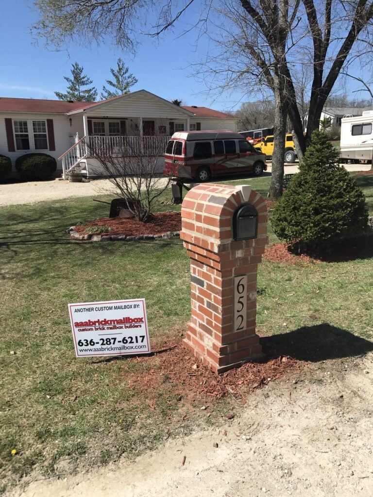 Arched red brick mailbox beside a gravel driveway, campers and work trucks parked in the yard behind