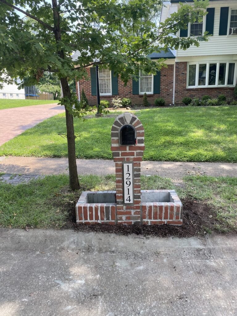 Arched red brick mailbox flanked by matching brick planter boxes, single tree beside the driveway