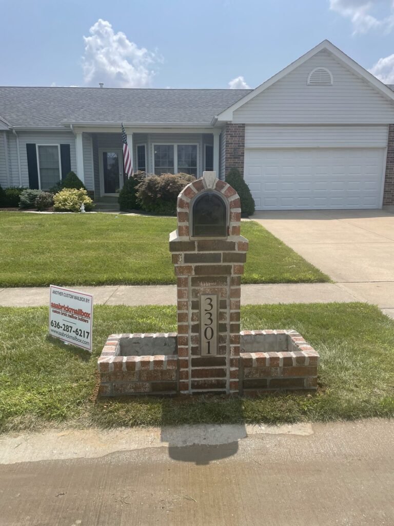 Arched brick mailbox flanked by two brick planter beds, ranch house with an American flag behind