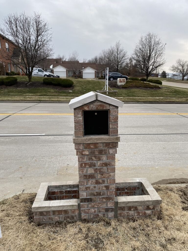 Square brick mailbox with a peaked stone cap and dark open mailbox slot, flanking planter beds still filled with bare soil