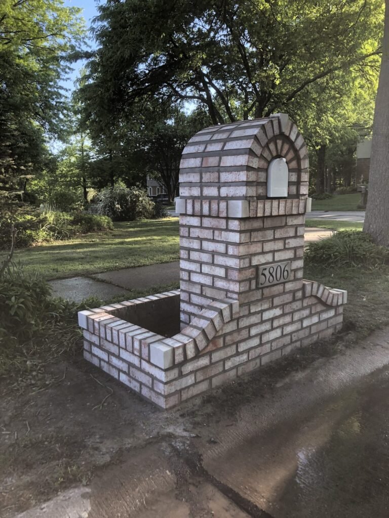 Rounded whitewashed brick mailbox with an L-shaped brick planter bed, viewed from the side