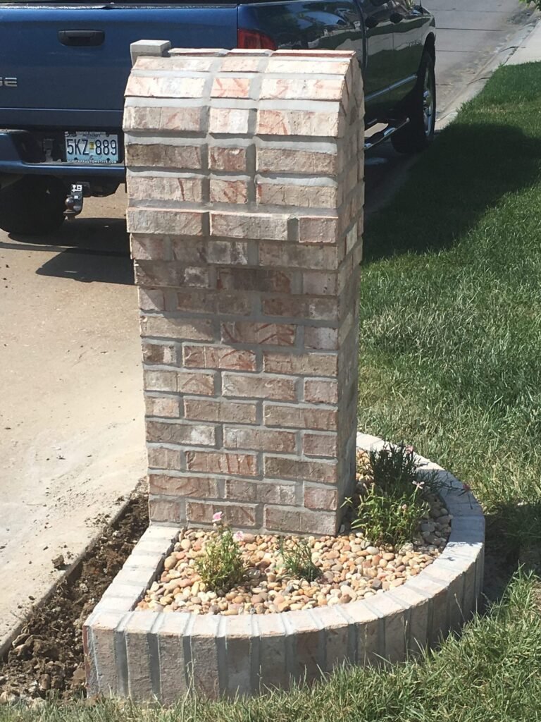 Close side view of a whitewashed brick mailbox column beside a curved planter bed with small flowering plants