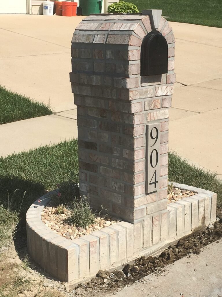 Arched brick mailbox in mixed gray and rust tones, set in a curved planter bed with river rock and small plants