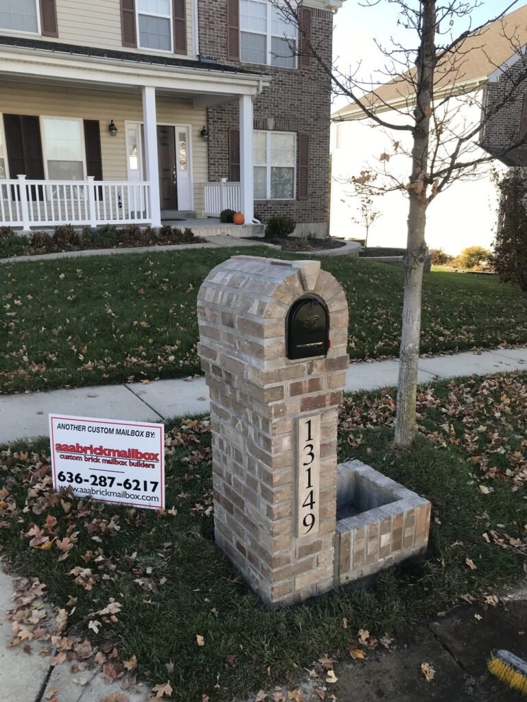 Arched brick mailbox with a single brick planter box, young tree beside it, two-story brick house with covered porch behind