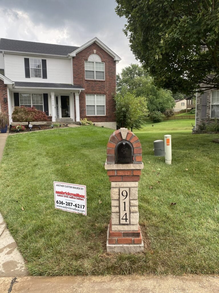 Arched red brick and gray stone dome top over a black mailbox, tan stone pillar with a house number plaque, on a corner lawn