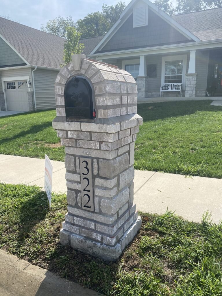 All light gray stacked stone mailbox with an arched dome top, stone number plate set into the pillar, near a sidewalk corner