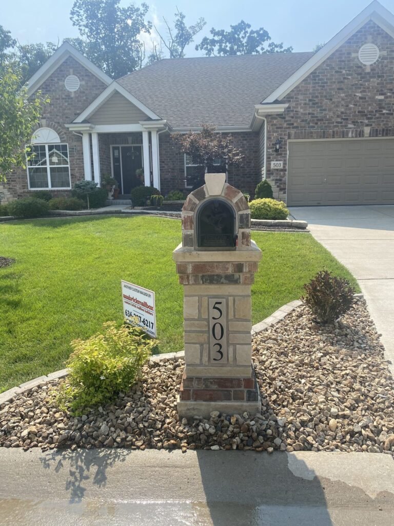 Reddish brick arched dome top on a stacked stone pillar with a number plaque, front view with driveway and shrub bed behind