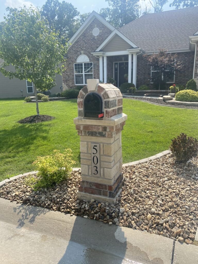 Same brick and stone arched mailbox seen from an angled side view near a young tree, number plaque on the pillar face