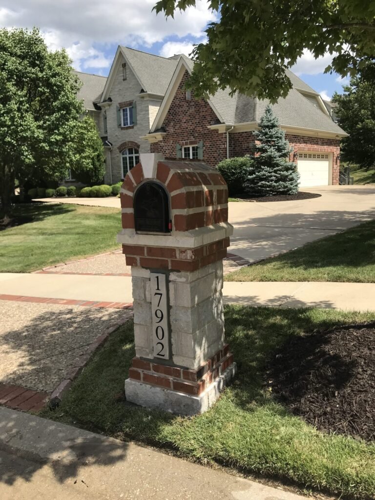 Wider view of a brick and stone arched mailbox under a tree canopy, with a two-story stone and brick home and garage behind it