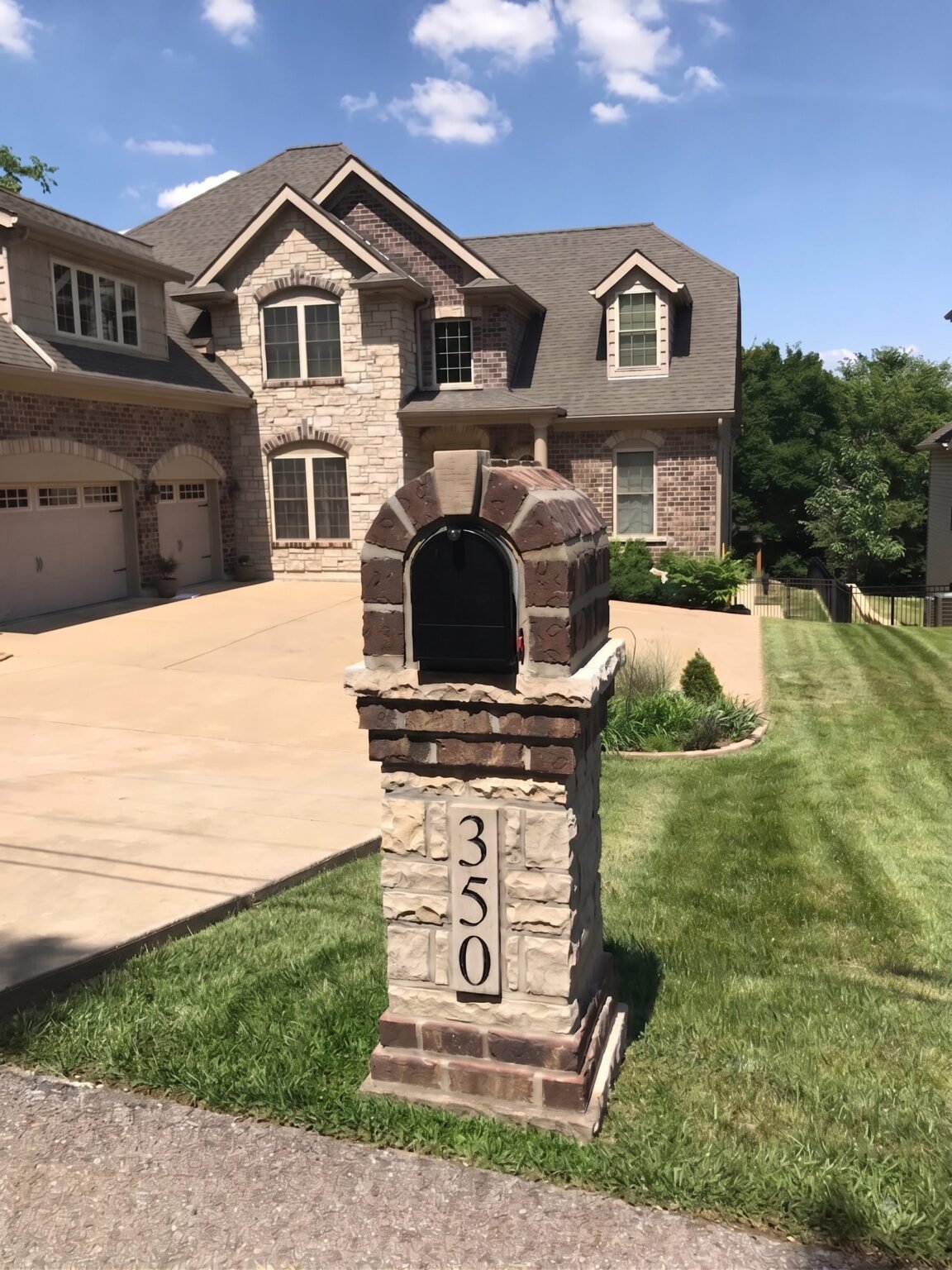 Brown and tan stacked stone mailbox with an arched dome top and number plaque, standing in front of a two-story brick and stone home