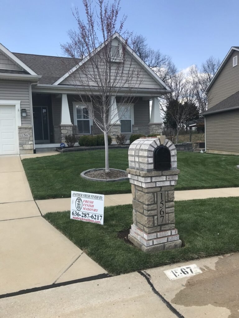 Multi-tone gray stacked stone mailbox with an arched dome top, number plate on the front face, young bare tree planted beside it