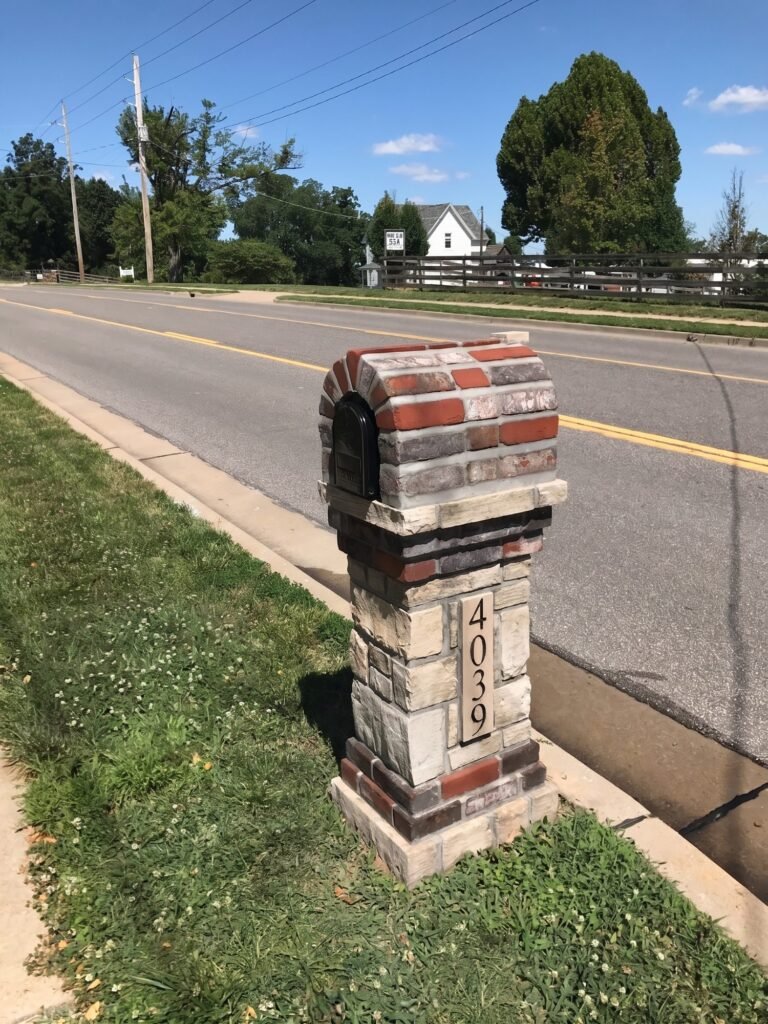 Red brick and gray stone banded arched top on a tan stacked stone pillar, number plate, standing roadside along a two-lane street