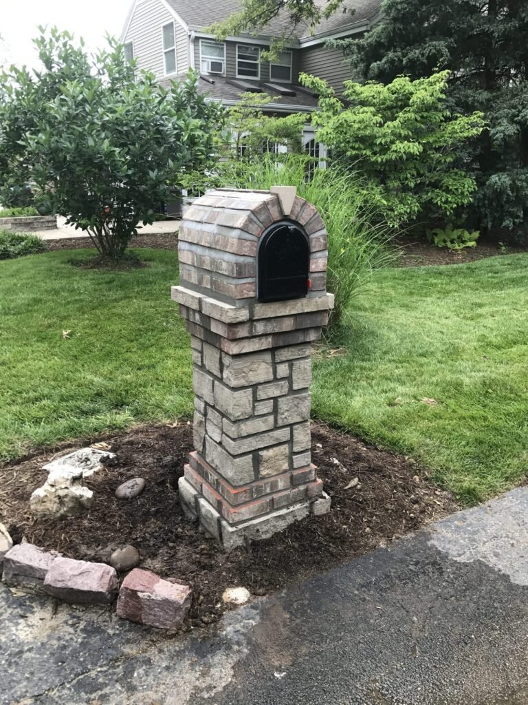 Rust and gray stacked stone mailbox with an arched dome top, set in a mulch bed with ornamental grasses and shrubs