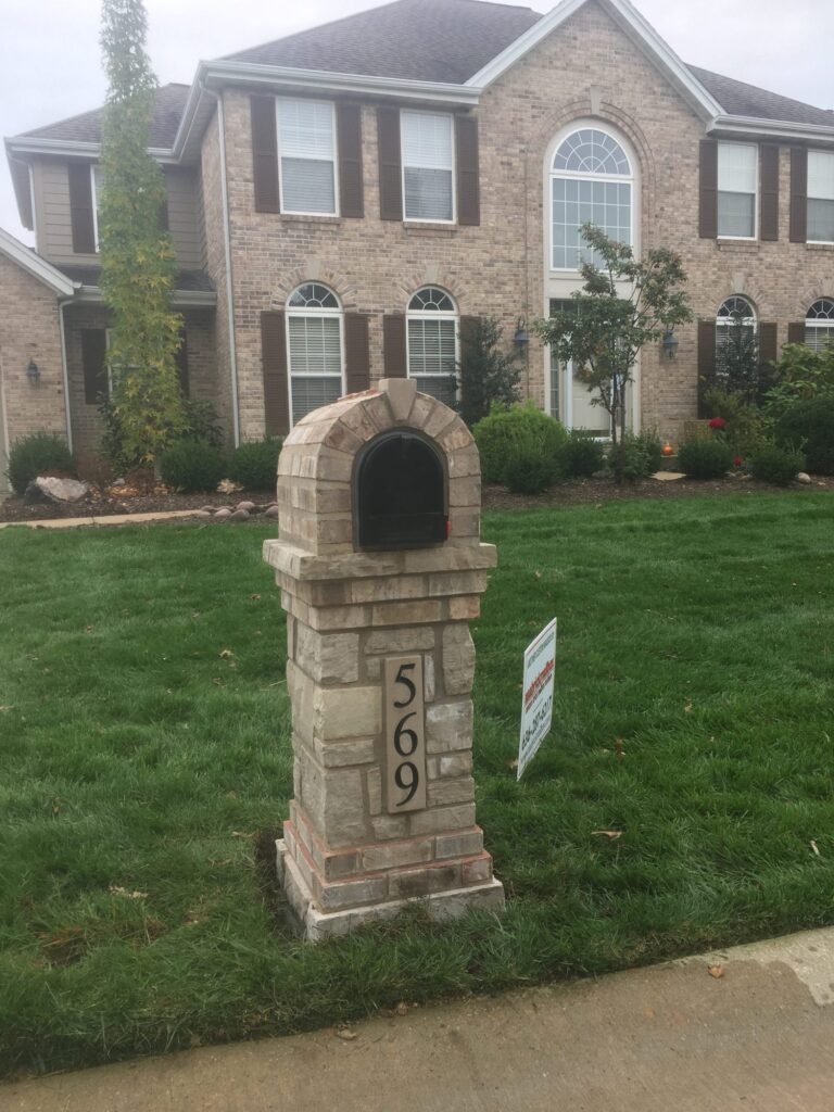 Tan stacked stone mailbox with an arched dome top and number plate, standing in front of a two-story brick home with dark shutters