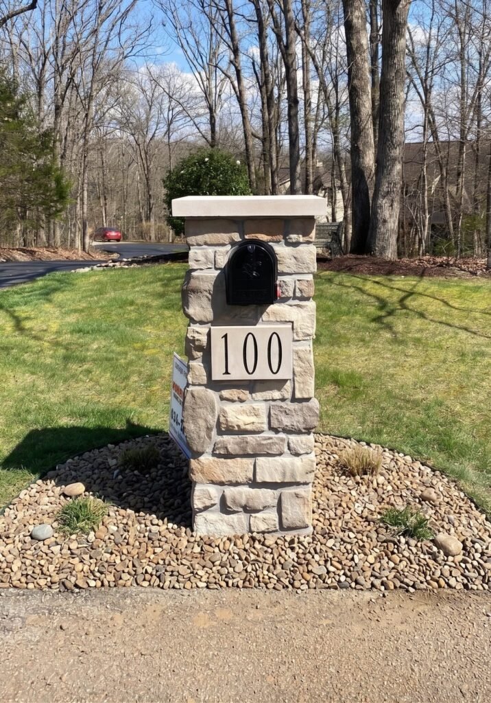 Flat stone slab cap on a tan stacked stone pillar with a number plate, set in a river-rock mulch bed along a wooded curve