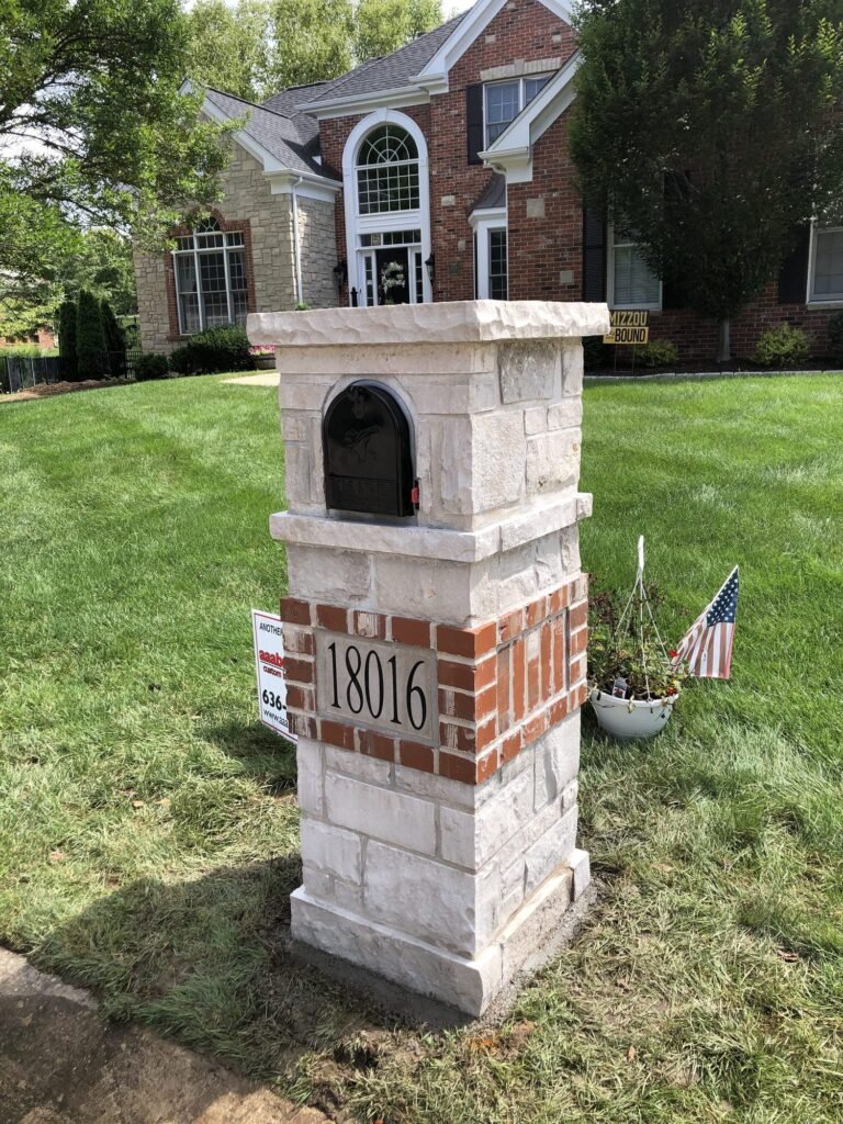 Arched brick dome top banded with tan stone, number plaque below the mailbox, hanging flower basket and American flag planted beside it