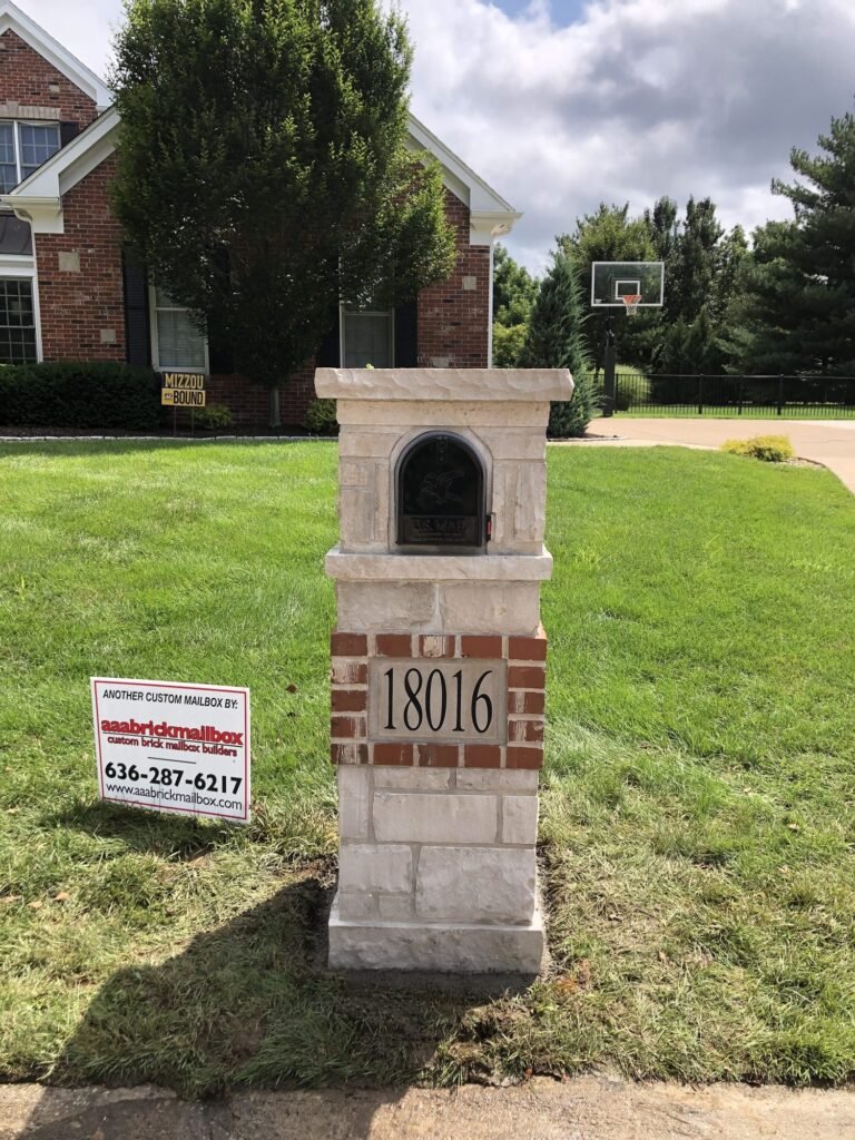 Closer view of the same arched brick and stone mailbox, number plaque visible, evergreen shrubs and a basketball hoop in the background