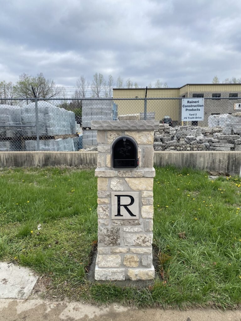 Flat stone cap over a light stacked stone pillar with a single letter plaque, standing in open grass with no house in view