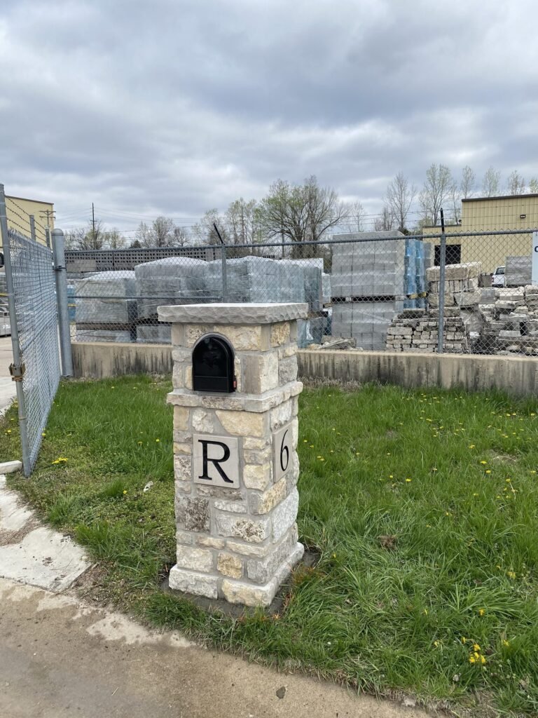 Same stone mailbox from another angle, showing a letter plaque paired with a single digit plaque, chain-link fence and stacked pavers behind