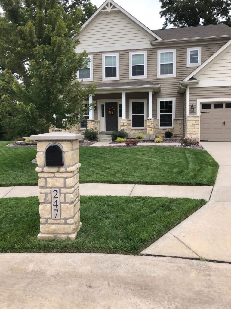 Flat stone slab cap on a tan stacked stone pillar with a number plate, standing in a mowed lawn beside a curving sidewalk