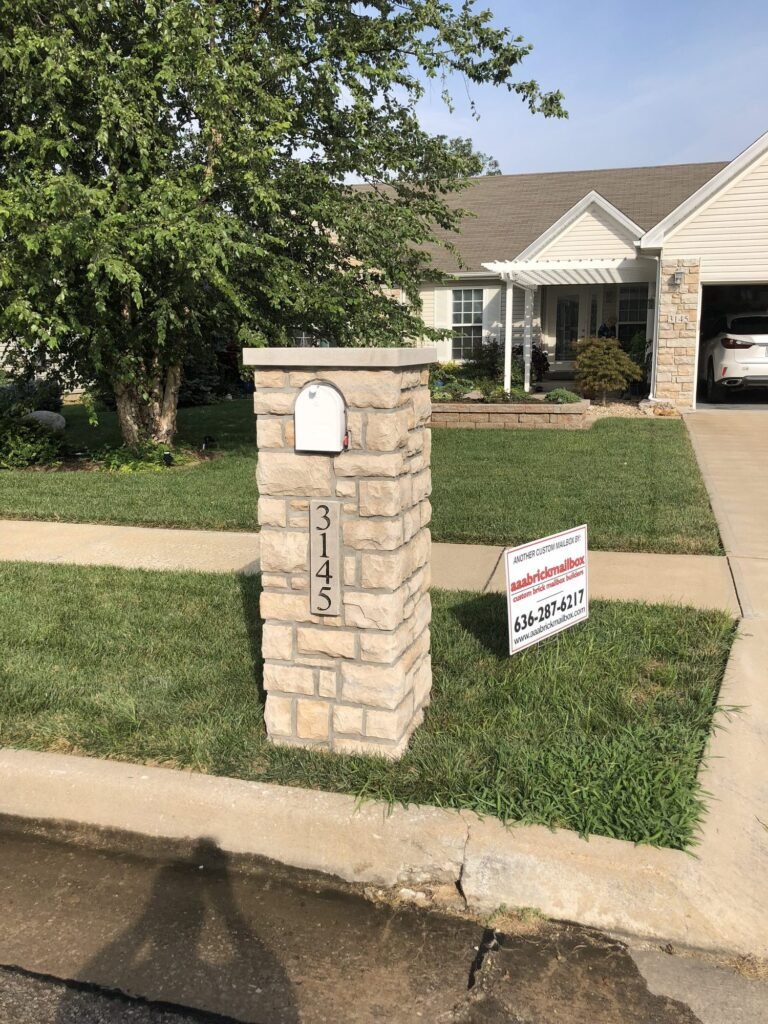Flat stone cap on a tan stacked stone pillar with a white mailbox door and number plate, ranch home with a pergola-covered porch behind