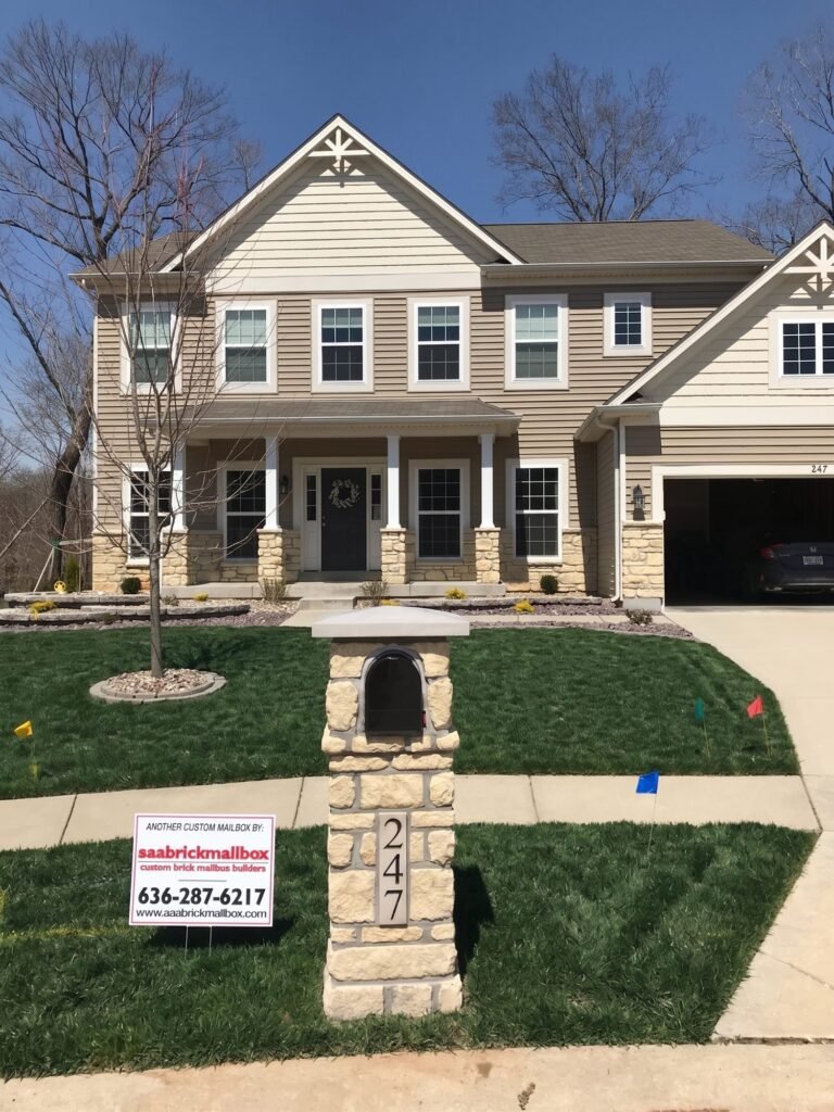 Closer view of the flat-capped tan stone mailbox and number plate, two-story home with stone porch columns and a car in the driveway