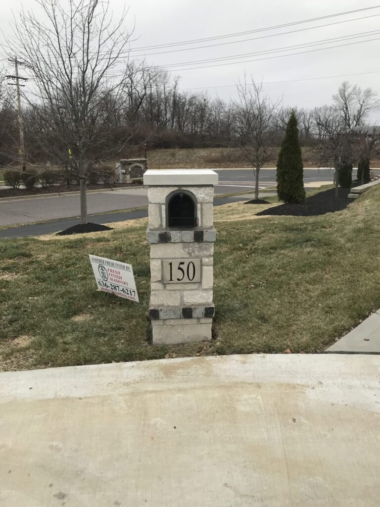 Flat stone cap on a light gray stone pillar banded with dark accent brick, number plate, standing roadside near young evergreen shrubs