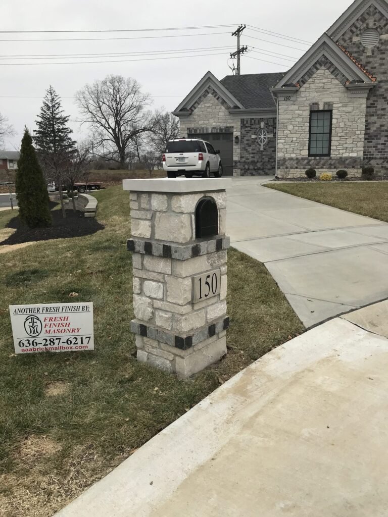 Same light gray stone mailbox with dark brick banding, wider view showing a stone-faced house and a parked SUV in the driveway