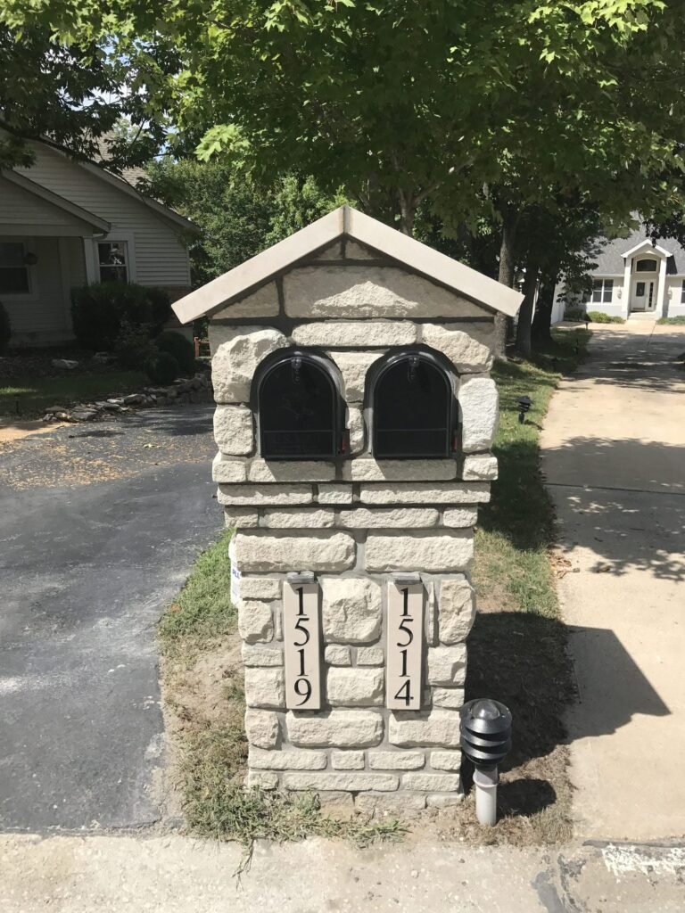 Twin mailboxes side by side under a peaked gable roof cap on a tan stacked stone pillar, two separate number plaques, shaded driveway entrance