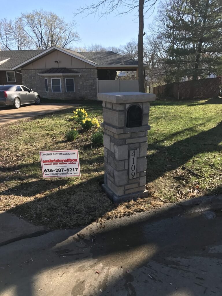Arched dome top on a tan stacked stone pillar with a number plate, daffodils blooming beside it in front of a brick ranch home