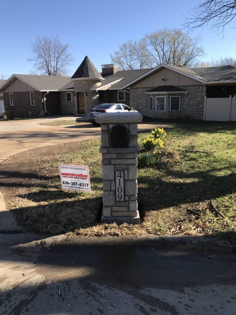 Gray stone mailbox with arched door and flat cap, yellow daffodils blooming beside it on a lawn