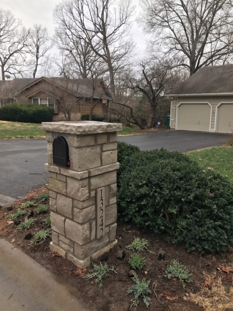 Flat-capped stone mailbox with black front-loading door and boxwood hedge beside a curved driveway