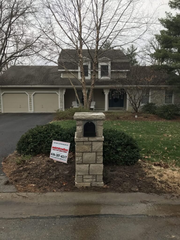 Tan stone mailbox with arched door and flat cap, flanked by trimmed boxwood hedges near a two-story home