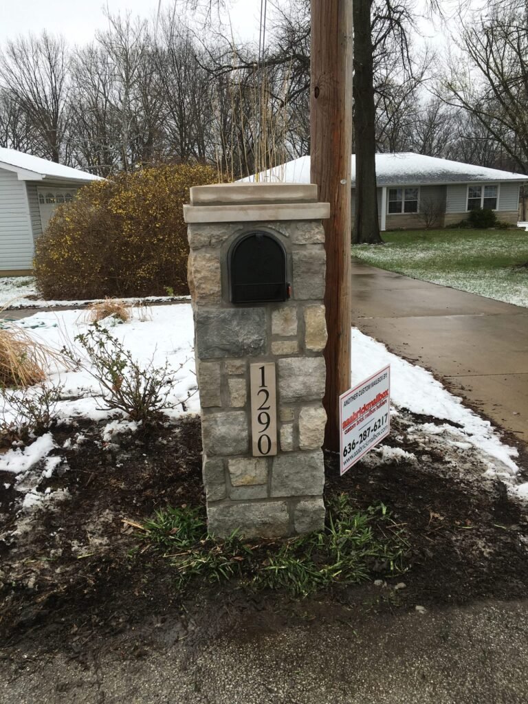 Mixed gray and tan stone mailbox with flat cap and arched door, standing in a light snow
