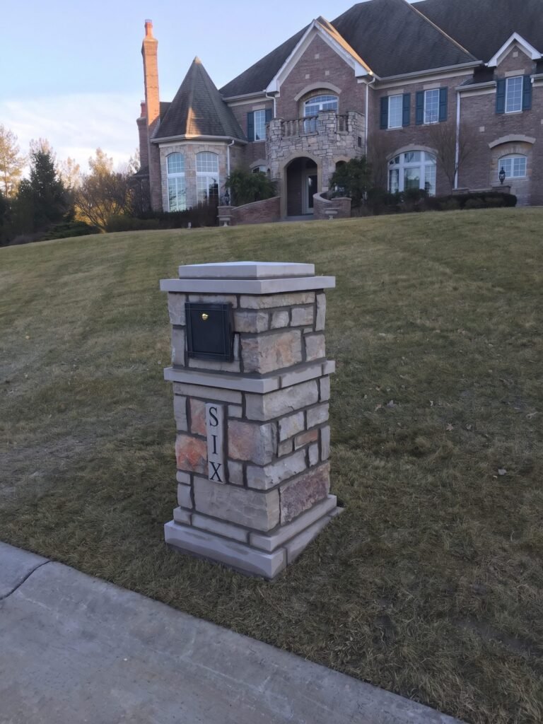 Gray stone mailbox with flat cap and front-loading door with a brass knob, large stone manor behind it