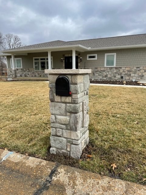 Gray stone mailbox with flat cap and arched door, seen from the side under an overcast sky