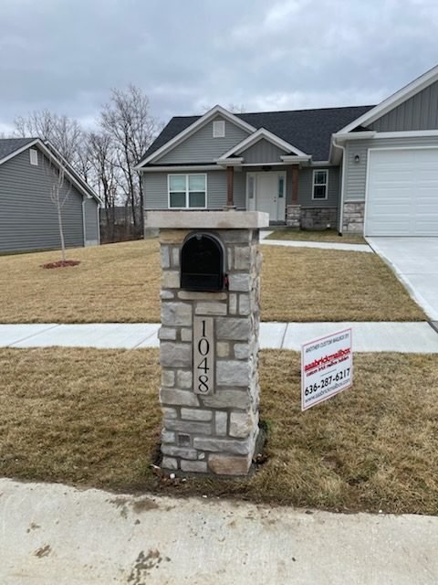 Gray fieldstone mailbox with a flat overhanging cap and arched door in front of a craftsman ranch home