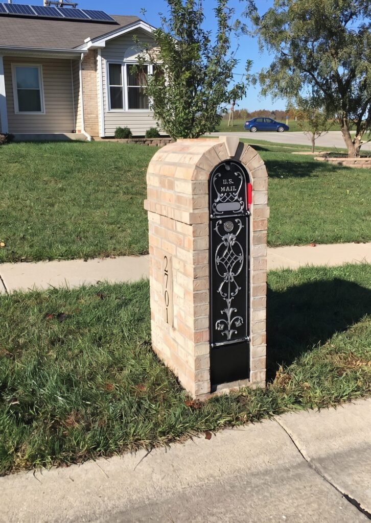 Tan brick mailbox with a barrel-vaulted arch top and ornate scrollwork door, solar panels visible on the roof
