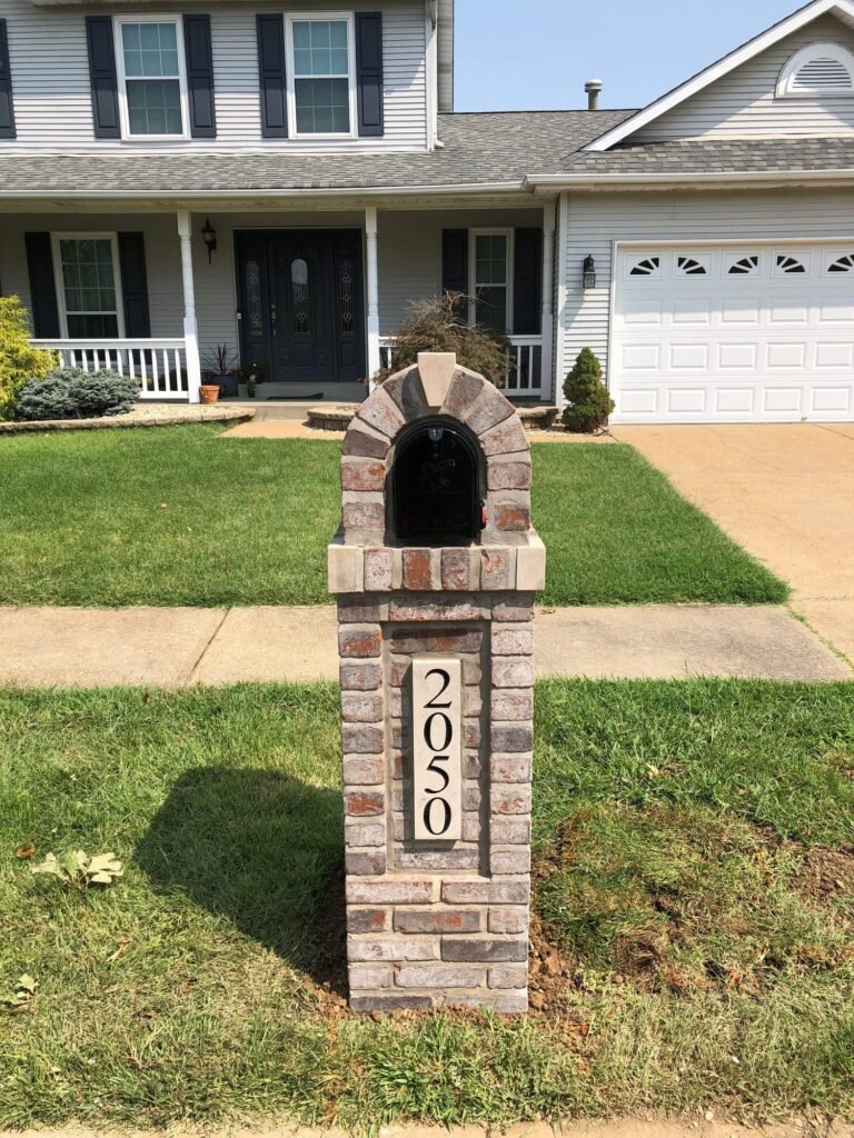 Brick mailbox with an arched top and tan keystone accent, two-story house with black shutters behind it