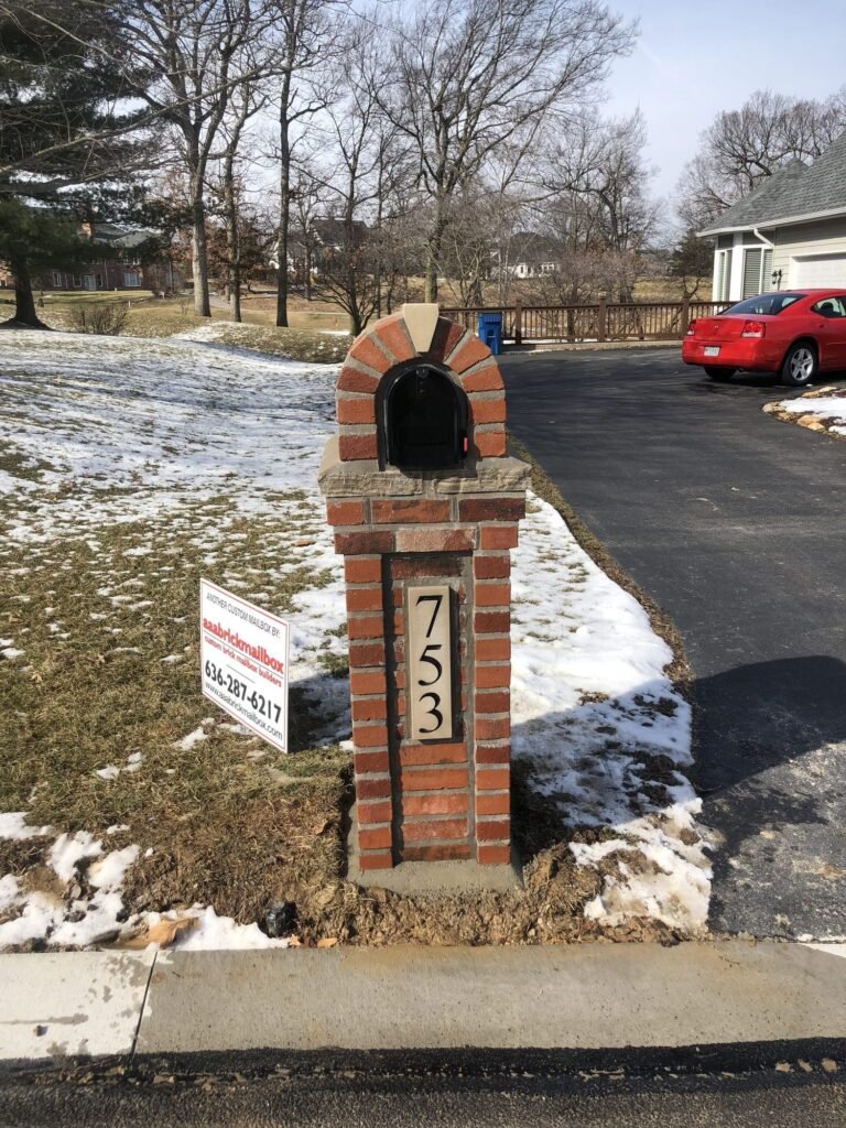Brick mailbox with an arched top and tan keystone accent, standing in patchy snow beside a driveway