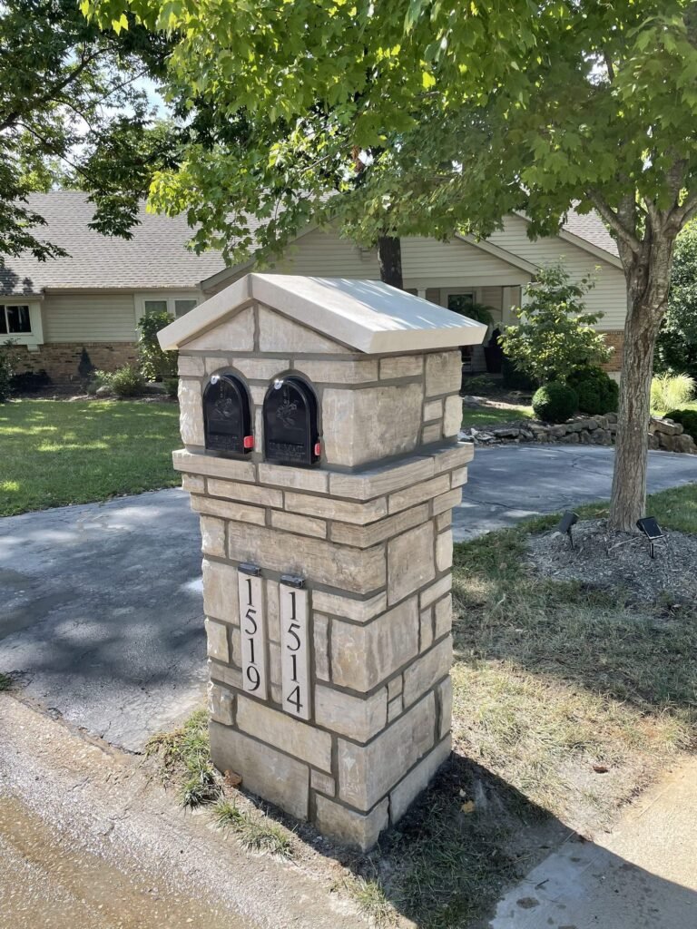 Double mailbox with a peaked stone roof and two arched doors, shaded by a tree over a driveway