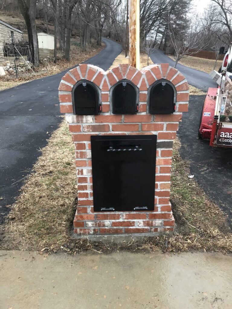 Triple mailbox with a scalloped triple-arch brick top and a hinged newspaper door, set along a gravel driveway