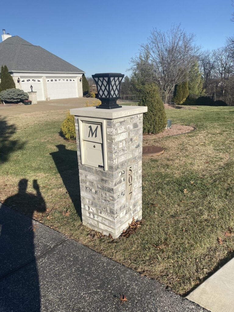 Brick mailbox with the same lantern-topped cap and monogram initial, seen from the side by a stone home