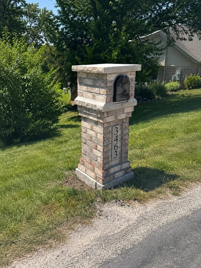 Tan and brown brick mailbox with a flat stone cap and bronze-toned door, set in a grassy yard