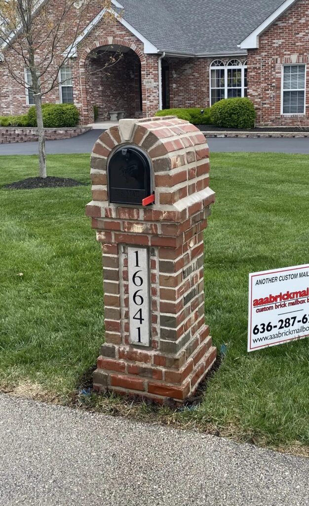 Arched brick mailbox in blended red and gray brick with a light stone cap, vertical stone address plaque, and a black arched door, brick ranch home behind with a yard sign nearby